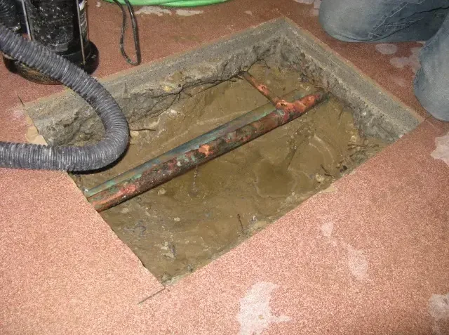Exposed copper pipe in a wet concrete floor opening with a vacuum hose resting beside the trench